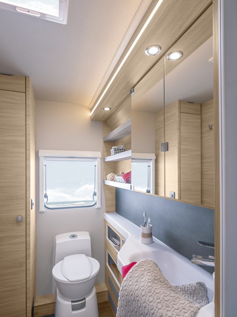 Bild från badrummet i husvagnen.  Längst bort en toalettstol med ett fönster ovanför. Till höger handfat och avställningsyta samt hyllor och spegelskåp.