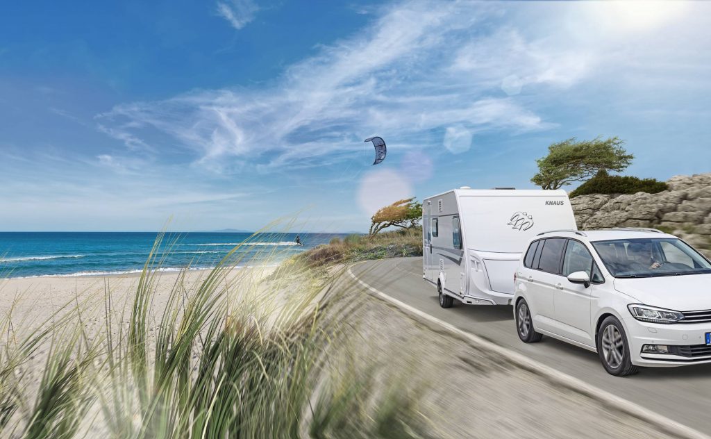 En vit bil med en husvagn kör på en väg längs med stranden, till vänster syns havet. Det är blå himmel och sommar.