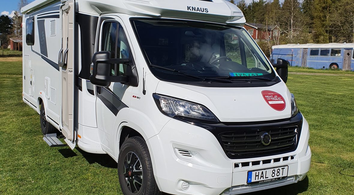 Vit husbil med Rent and Travel-dekal på motorhuven står på en grön gräsmatta. I bakgrunden syns träd, delar av ett rött hus, den blå himlen och några fluffiga, vita moln.